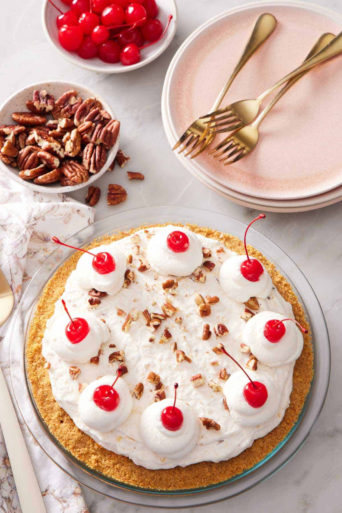 Overhead view of a Millionaire Pie with dollops of whipped cream topped with maraschino cherries. A stack of plates, forks, bowl of pecans, and more maraschino cherries off to the side.