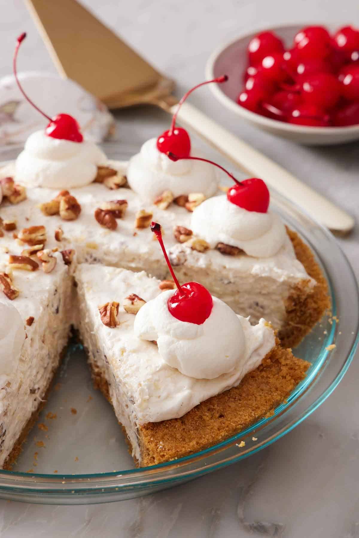 A slice of Millionaire Pie sliced in the pie dish, topped with chopped pecans, whipped cream, and maraschino cherries. A bowl of maraschino cherries in the background.