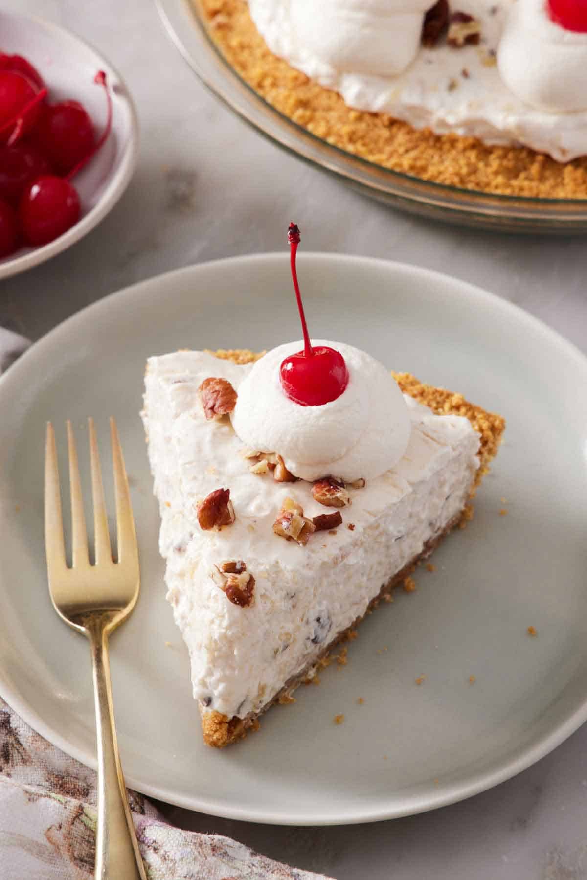 A plate with a slice of Millionaire Pie with a dollop of whipped cream with a maraschino cherry on top with a fork beside it.