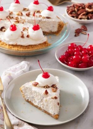A plate with a slice of Millionaire Pie with a dollop of whipped cream with a maraschino cherry on top. A bowl of more maraschino cherries in the back along with the rest of the pie.