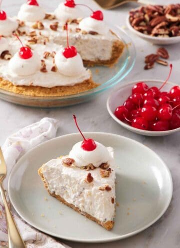 A plate with a slice of Millionaire Pie with a dollop of whipped cream with a maraschino cherry on top. A bowl of more maraschino cherries in the back along with the rest of the pie.