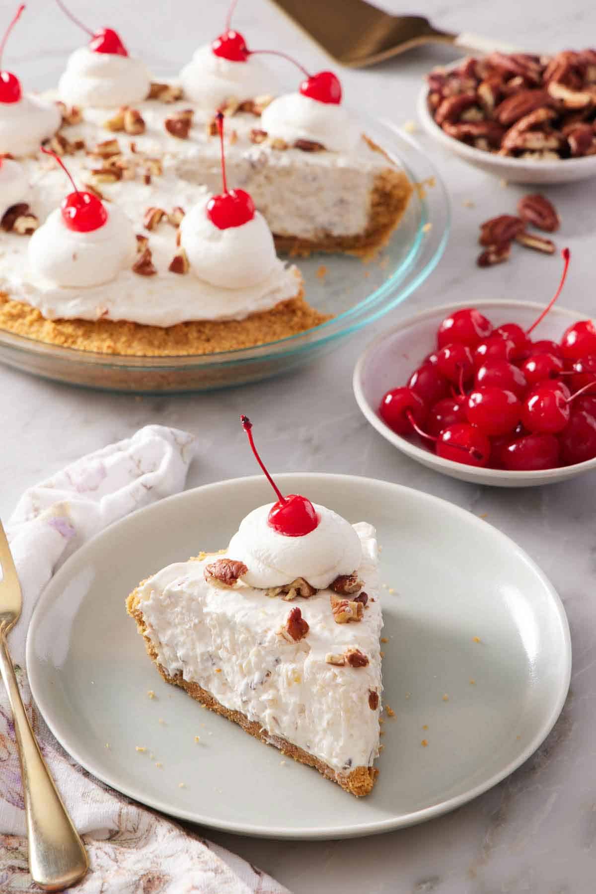 A plate with a slice of Millionaire Pie with a dollop of whipped cream with a maraschino cherry on top. A bowl of more maraschino cherries in the back along with the rest of the pie.