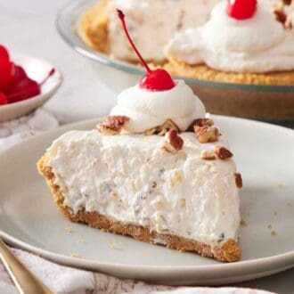 A side view of a plate with a slice of Millionaire Pie with a dollop of whipped cream with a maraschino cherry on top. A bowl of more maraschino cherries in the back with the rest of the pie.