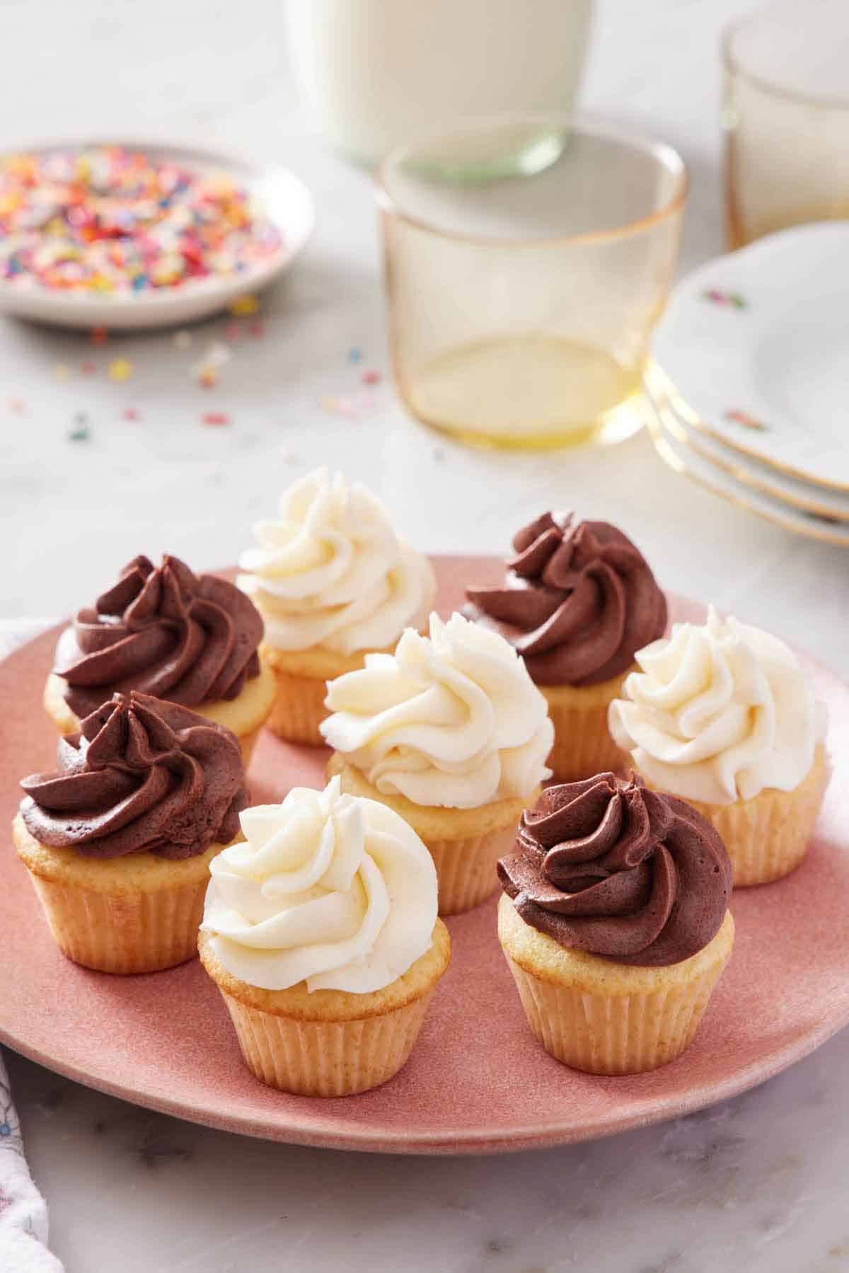 A pink plate with four mini cupcakes with chocolate frosting and four with vanilla frosting. A plate of sprinkles in the background along with glasses.