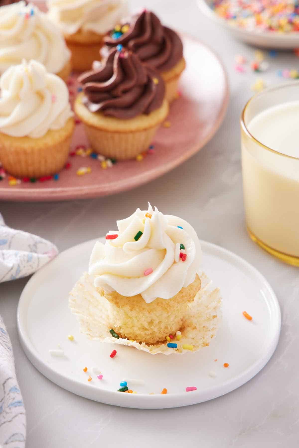 A plate with a mini cupcake with the paper liner pulled down and with sprinkles in the frosting. A glass of milk and more mini cupcakes in the background.