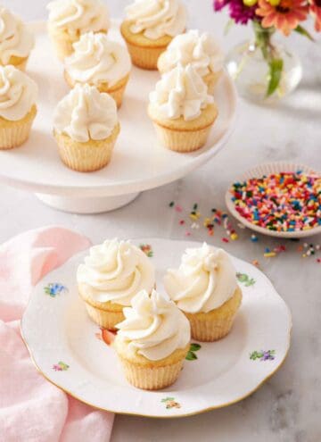 A plate with three mini cupcakes with a cake stand in the back with more cupcakes and a plate of sprinkles.