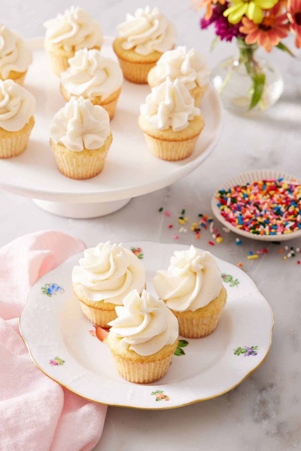 A plate with three mini cupcakes with a cake stand in the back with more cupcakes and a plate of sprinkles.