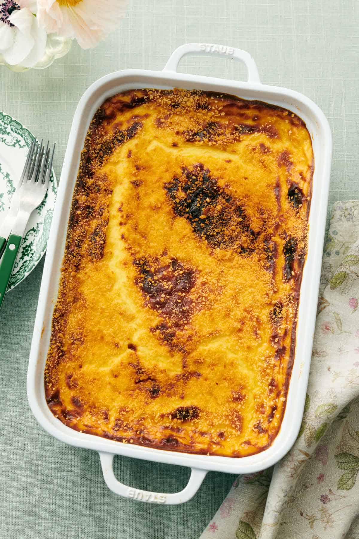 Overhead view of a white baking dish with a freshly baked moussaka. A napkin on the side along with some forks and plates.