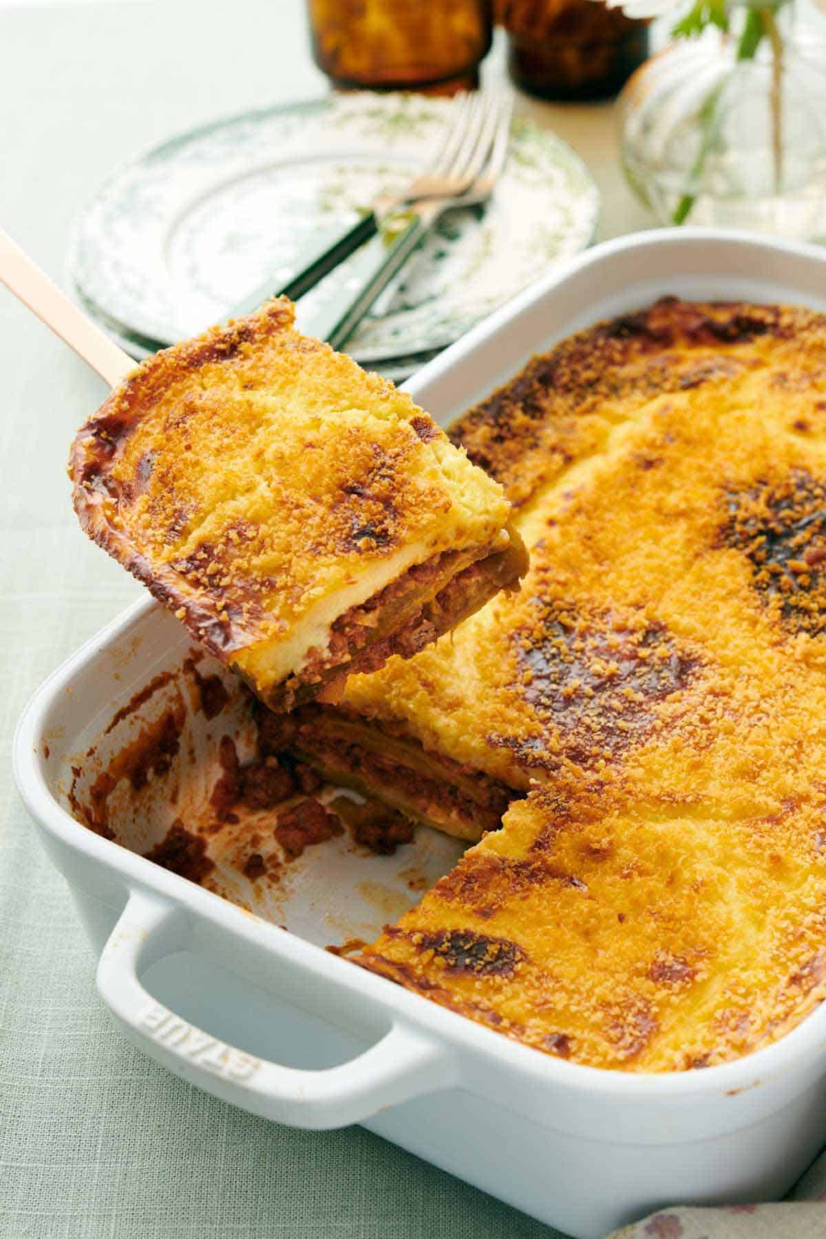 A piece of moussaka lifted from a baking dish. Plates and forks in the background.