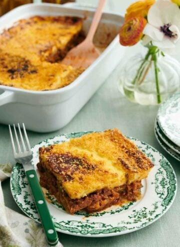 A piece of moussaka on a plate with a fork. A vase of flower and a baking dish with the rest of the moussaka in the background.