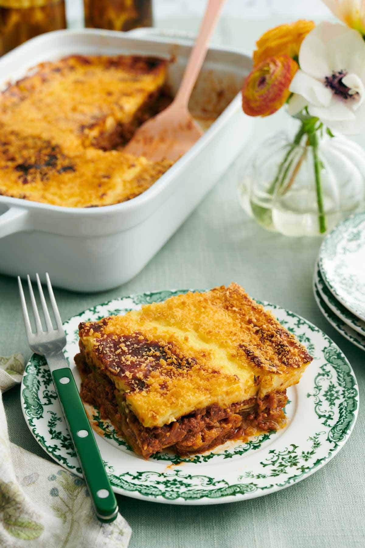 A piece of moussaka on a plate with a fork. A vase of flower and a baking dish with the rest of the moussaka in the background.