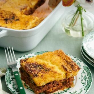 Pinterest graphic of a serving of moussaka on a plate with a fork. A vase of flower and a baking dish with the rest of the moussaka in the background.