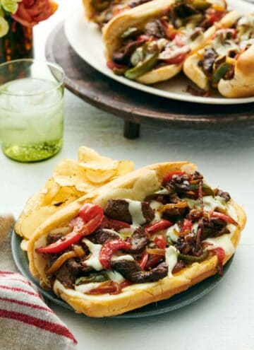 A Philly cheesesteak on a plate along with chips. A drink in the back along with a platter of three more Philly cheesesteaks.