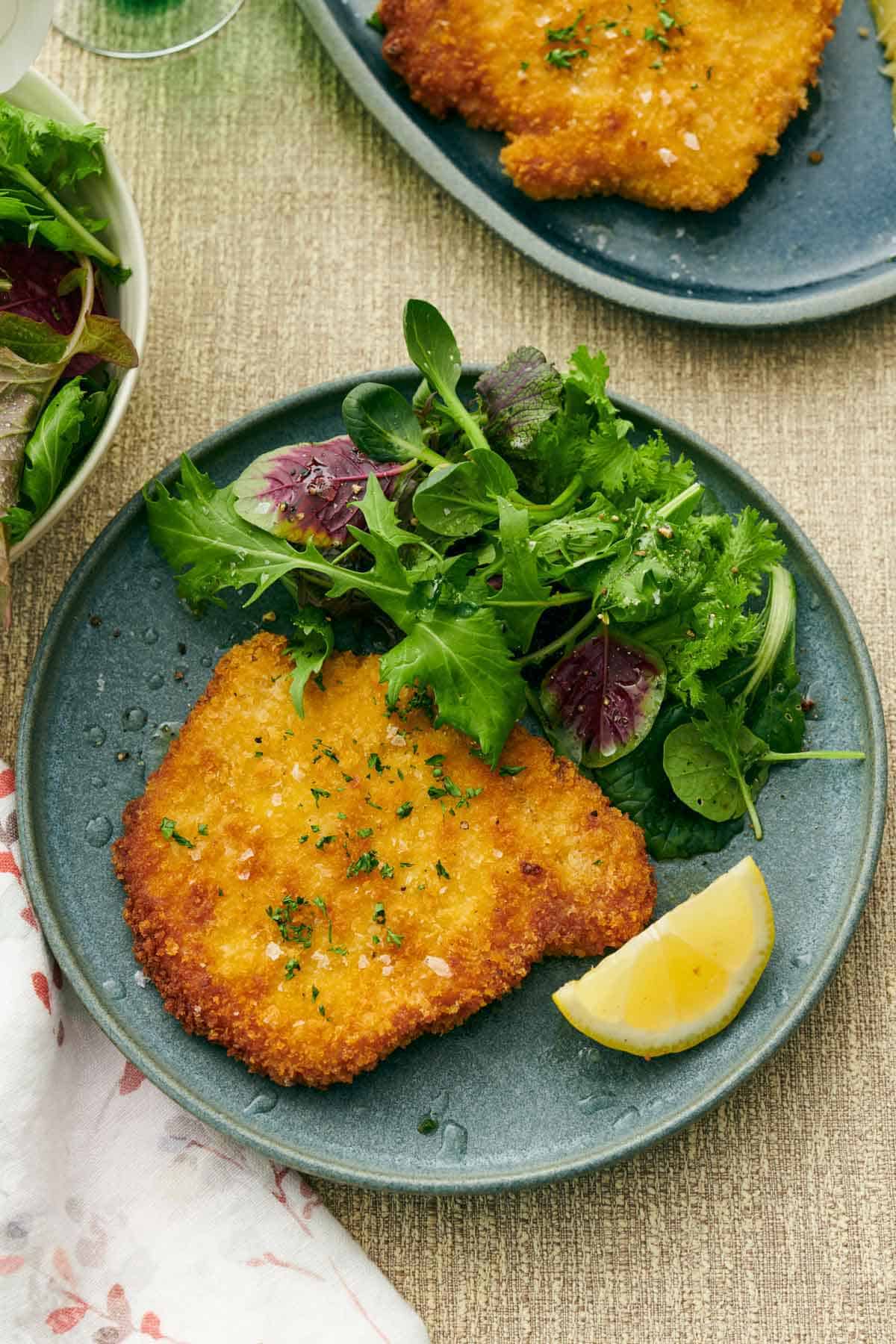 Overhead view of a plate with a schnitzel, mixed greens, and a lemon wedge. Chopped parsley and flaky salt garnished on top.