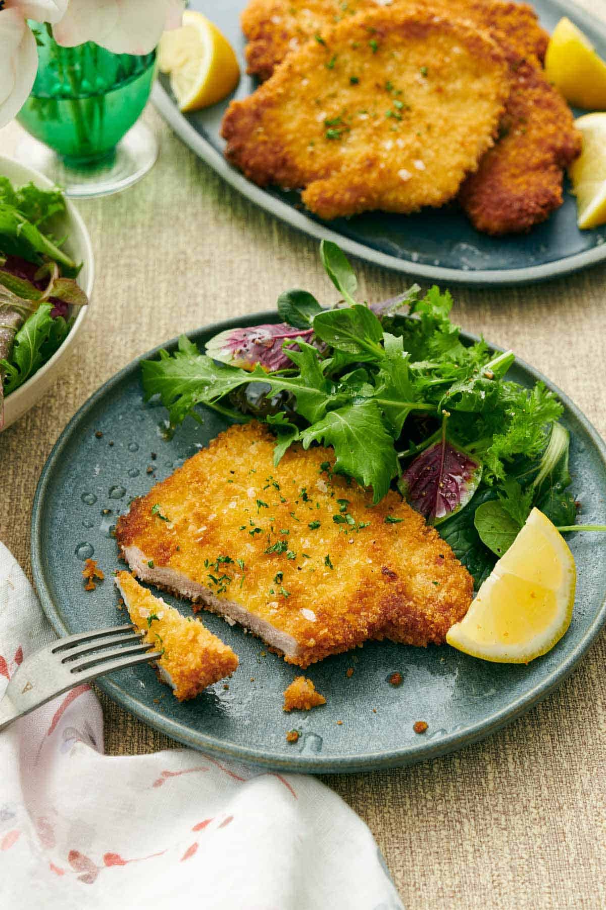 A plate with a schnitzel cut with a bite on a fork. Mixed greens and a lemon wedge beside it. More pork schnitzels in the background.