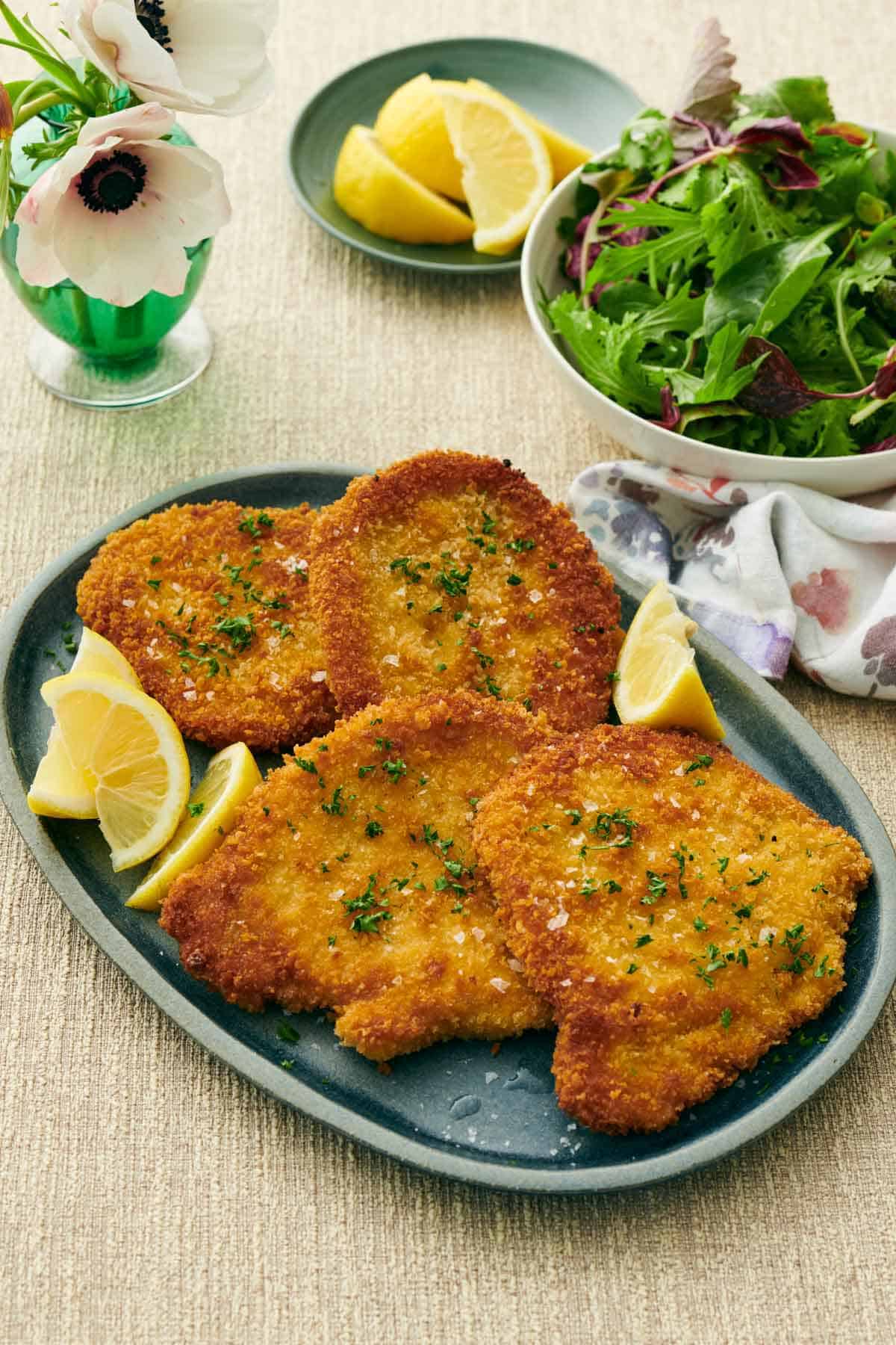 A platter of four pork schnitzels with parsley and flaky salt garnished on top surrounded by lemon wedges. A bowl of mixed greens and more lemon wedges in the background.