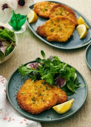 A plate with a crispy schnitzel, mixed greens, and lemon wedge. A platter with more pork schnitzels in the background along with more lemon wedges. A flower off to the side.