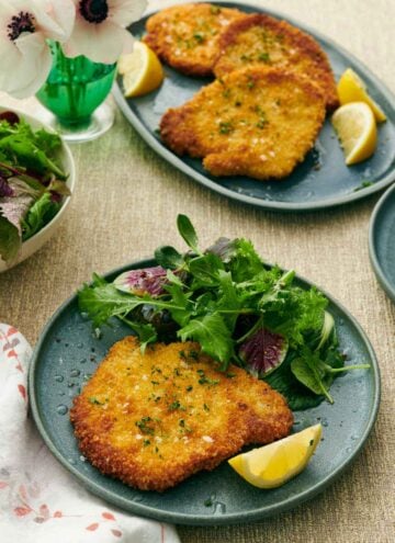 A plate with a crispy schnitzel, mixed greens, and lemon wedge. A platter with more pork schnitzels in the background along with more lemon wedges. A flower off to the side.