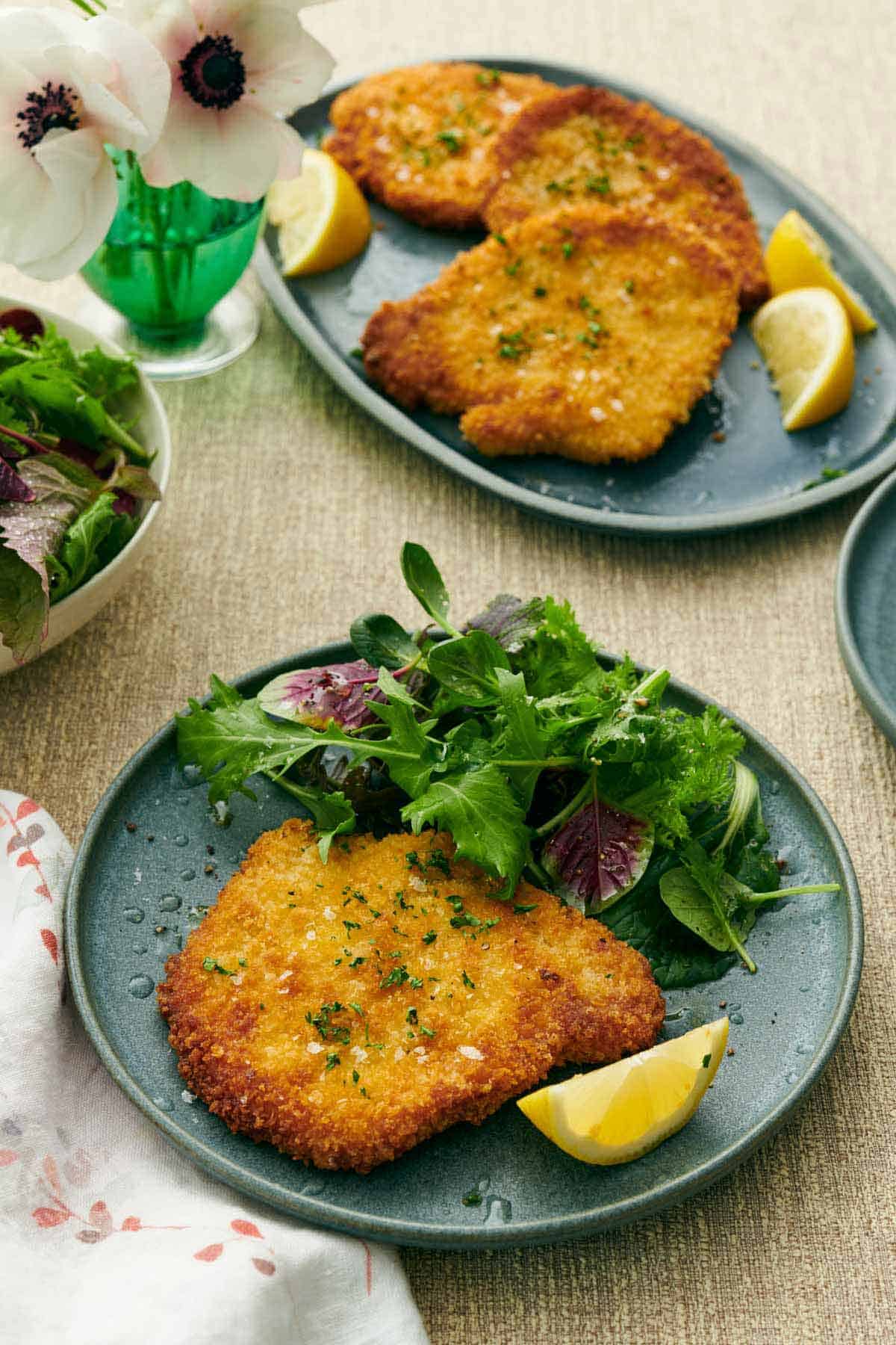 A plate with a crispy schnitzel, mixed greens, and lemon wedge. A platter with more pork schnitzels in the background along with more lemon wedges. A flower off to the side.