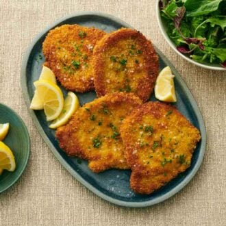 An overhead view of a platter of schnitzels topped with salt and parsley surrounded by lemon wedges. A bowl of lemon wedges and mixed greens on the side.