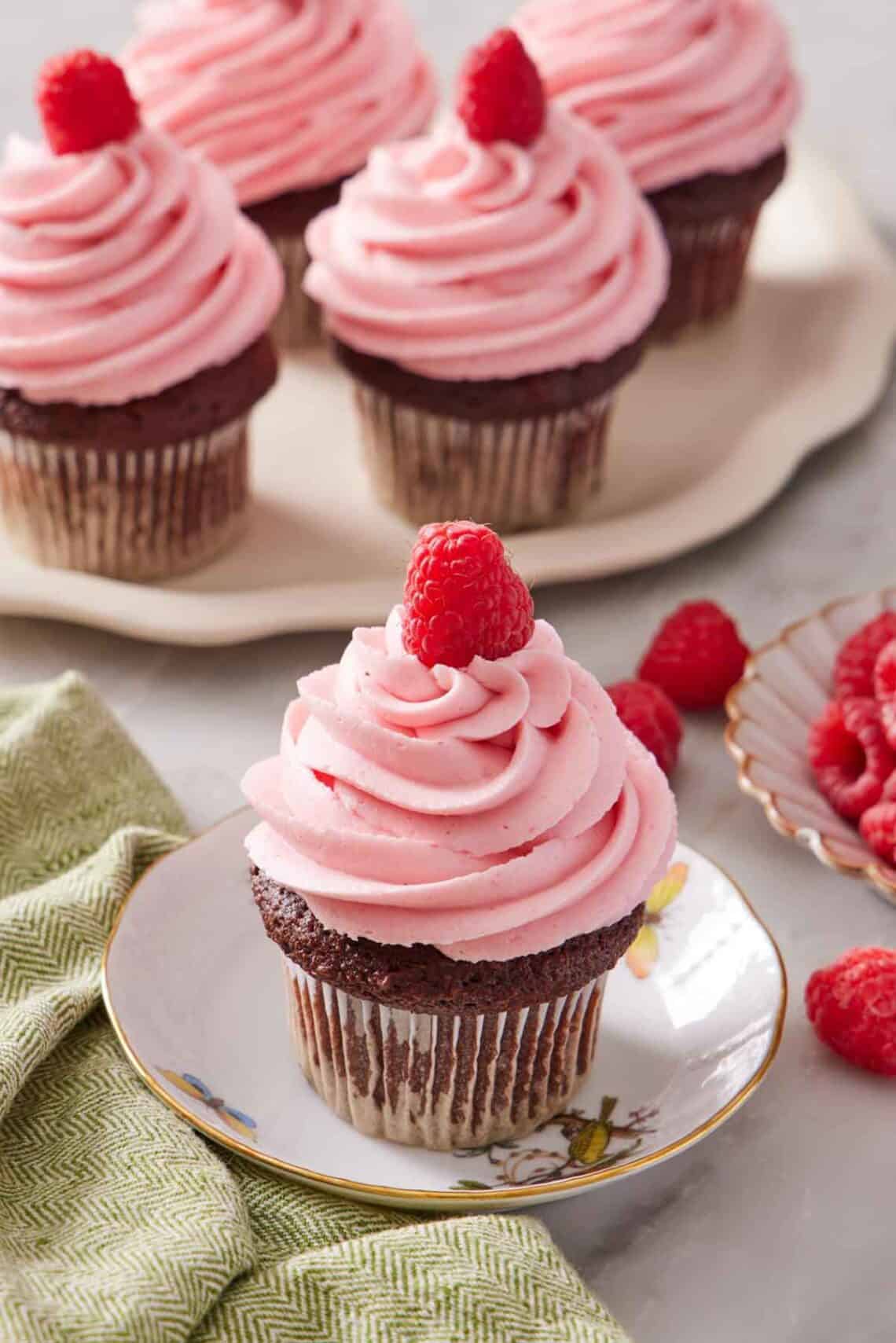 A plate with a cupcake topped with raspberry buttercream frosting and a fresh raspberry. More frosted cupcakes in the background. Fresh raspberries scattered on the surface.