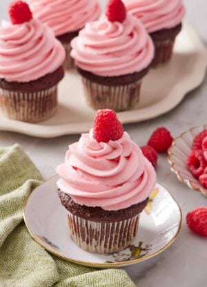 A plate with a cupcake topped with raspberry buttercream frosting and a fresh raspberry. More frosted cupcakes in the background. Fresh raspberries scattered on the surface.