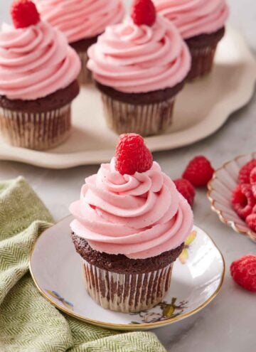 A plate with a cupcake topped with raspberry buttercream frosting and a fresh raspberry. More frosted cupcakes in the background. Fresh raspberries scattered on the surface.