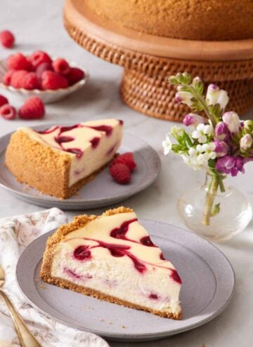 Two plates with slices of raspberry cheesecake. A mini vase of flowers with the rest of the cake in the background along with fresh raspberries.