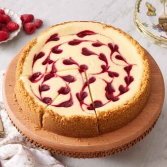 A raspberry cheesecake on a cake stand with two slices cut. A stack of plates and forks along with a plate of raspberries in the background.