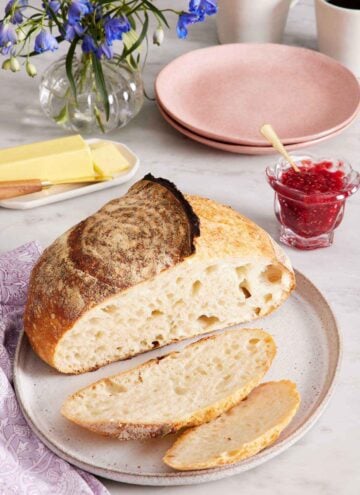 A plate with a loaf of sourdough bread with two slices cut off in front. A jar of jam, plates, butter, flowers, and two cups of coffee in the background.