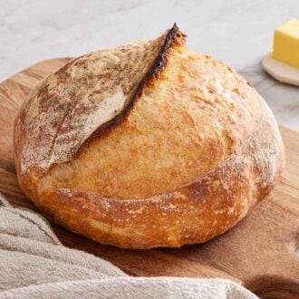 A wooden serving board with a loaf of sourdough bread with a linen napkin. A stick of butter in the back.