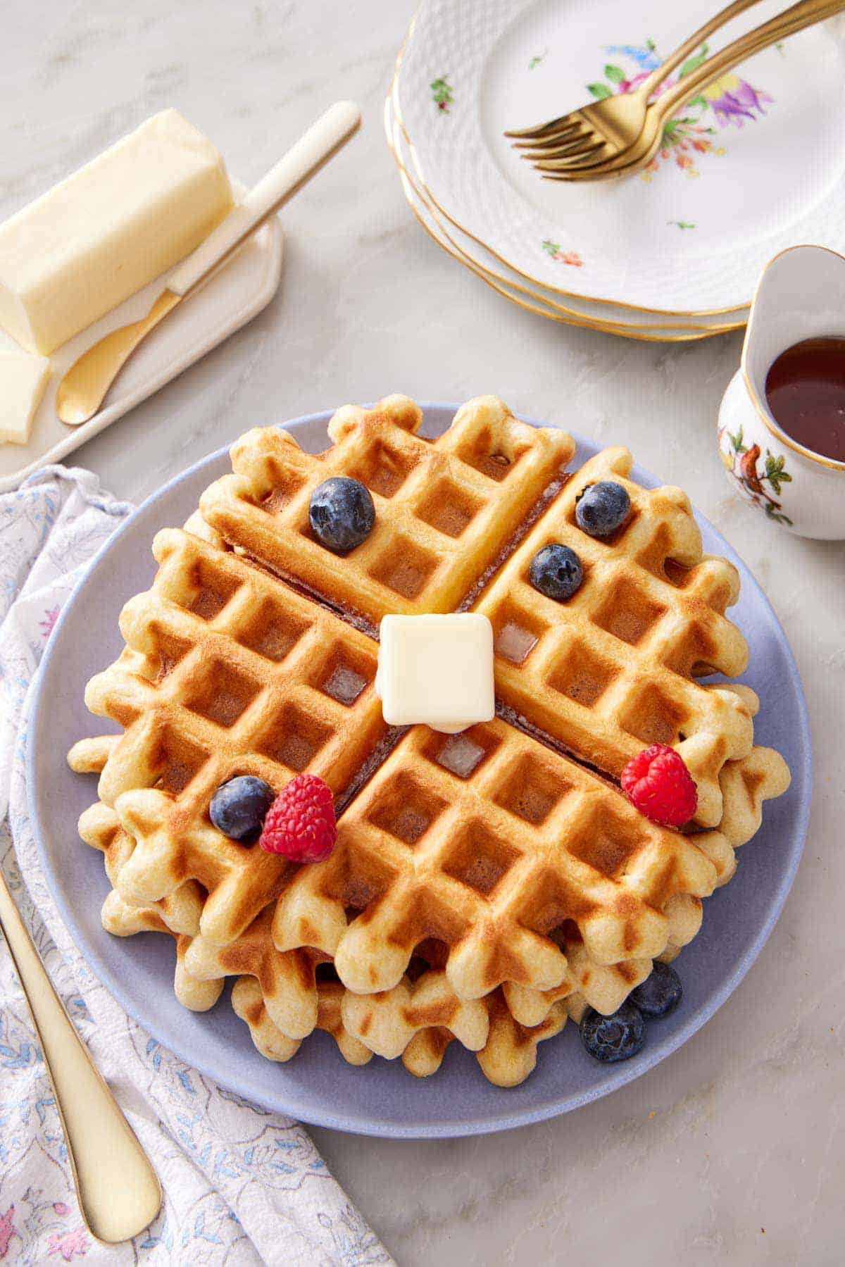 A plate with a stack of sourdough waffles with syrup, butter, and berries on top. Butter, syrup, stack of plates, and forks off to the side.