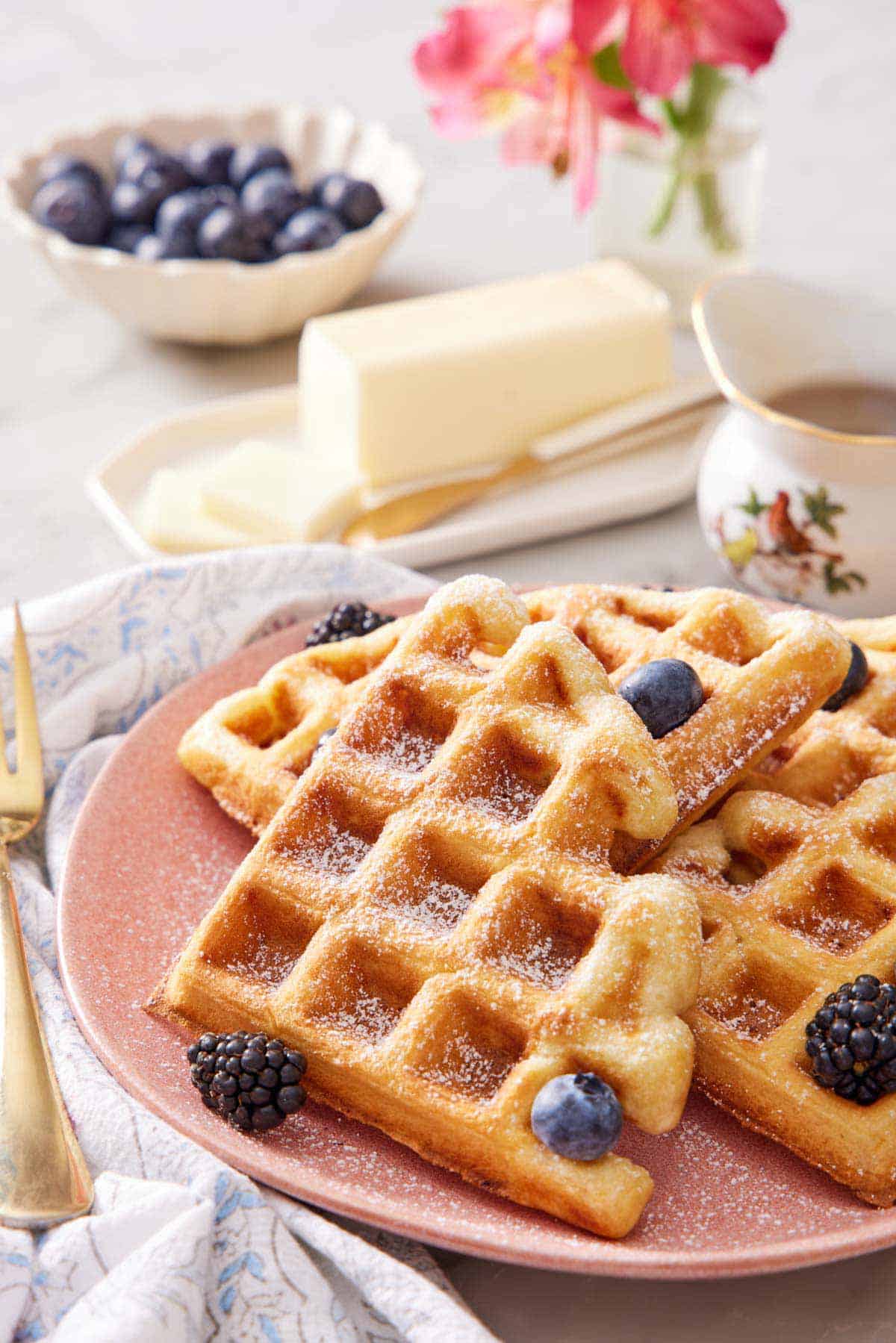 A plate with cut sourdough waffles topped with powdered sugar and berries. Butter in the background along with a bowl of berries and syrup.