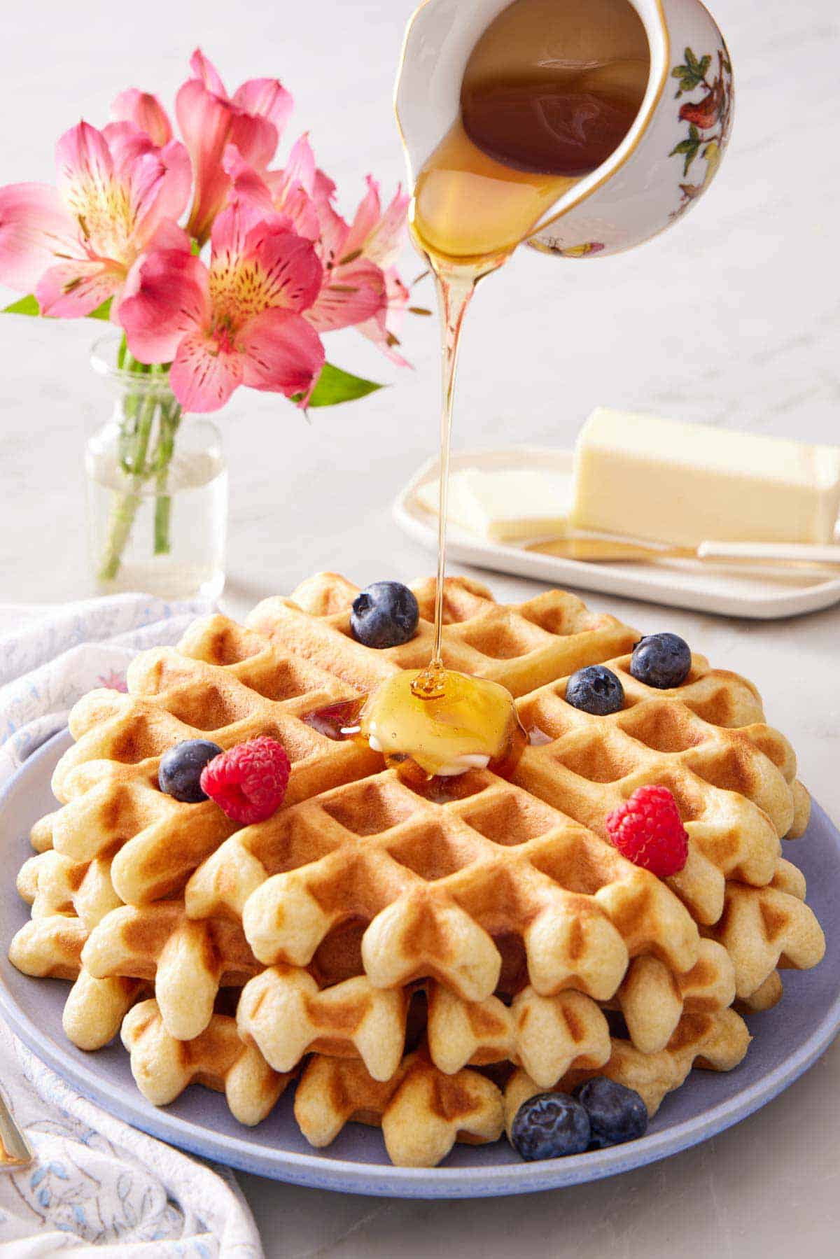 Maple syrup poured over a plate of sourdough waffles with berries. Flowers in the background with a stick of butter.