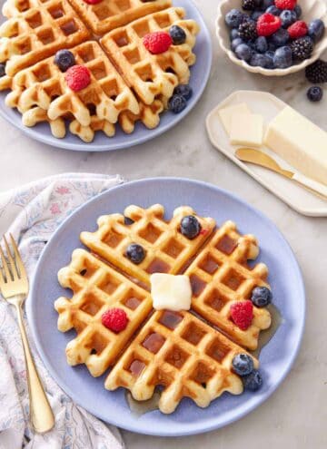 A purple plate with a sourdough waffle topped with syrup, butter, and berries. Another plate of waffles and berries in the back along with a bowl of berries and butter.