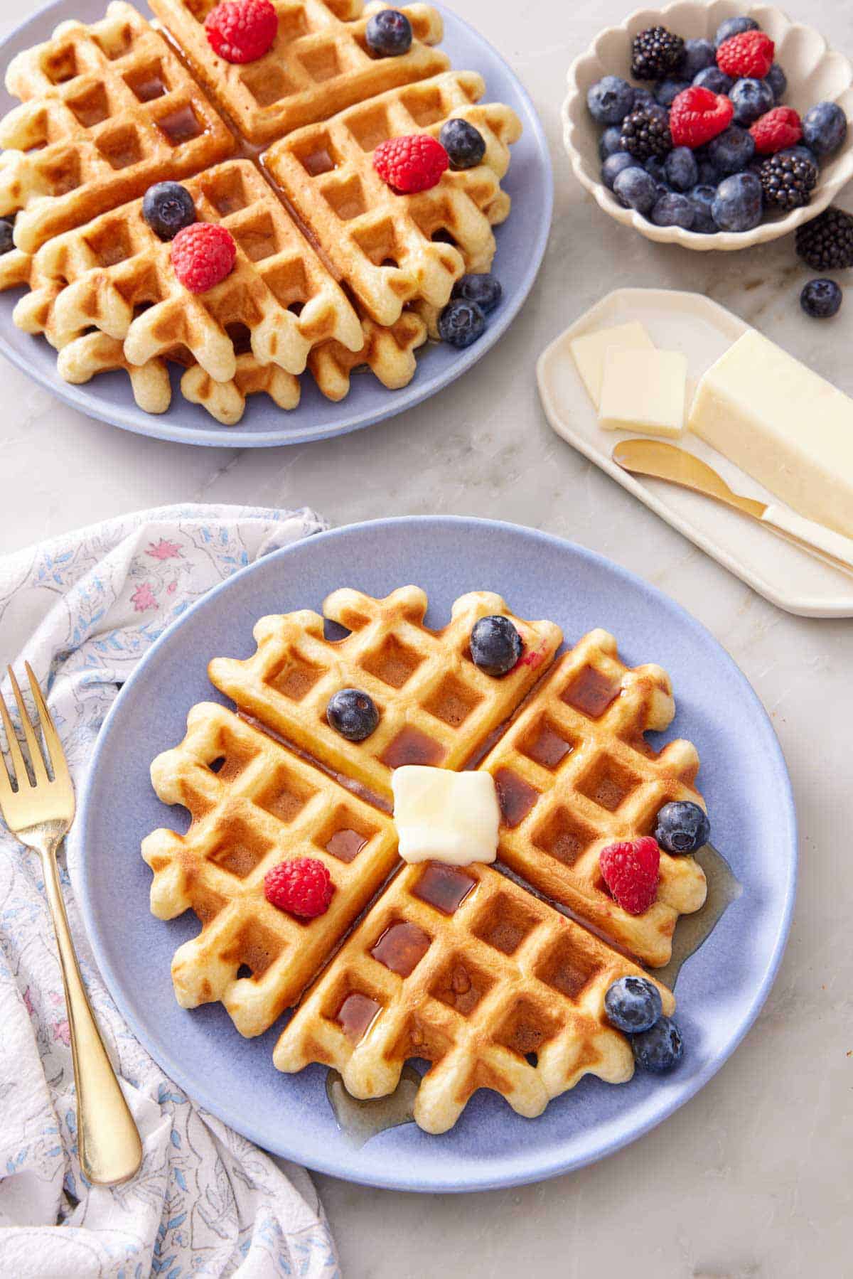 A purple plate with a sourdough waffle topped with syrup, butter, and berries. Another plate of waffles and berries in the back along with a bowl of berries and butter.