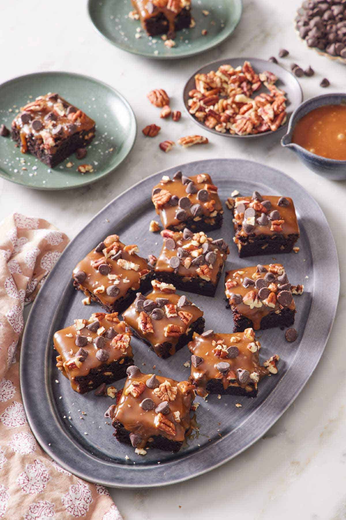 A platter of turtle brownies with two plated servings in the back along with a plate of pecans and bowl of caramel sauce.