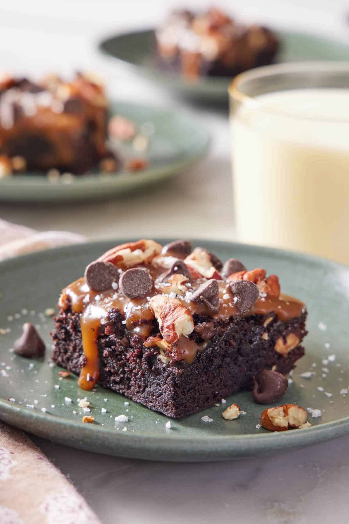 A plate with a piece of turtle brownies with flaky salt sprinkled on top. A glass of milk and more brownies in the background.