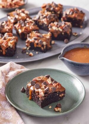 A plate with a piece of turtle brownies with a platter more in the background along with a bowl of caramel.