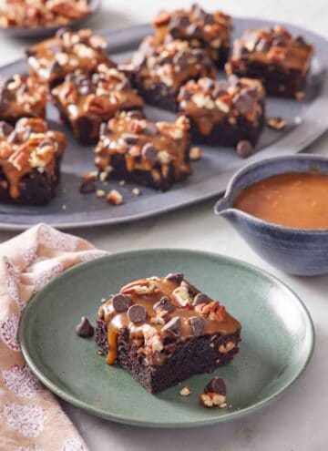 A plate with a piece of turtle brownies with a platter more in the background along with a bowl of caramel.