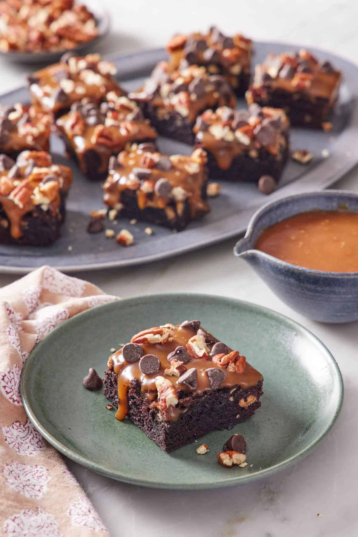 A plate with a piece of turtle brownies with a platter more in the background along with a bowl of caramel.