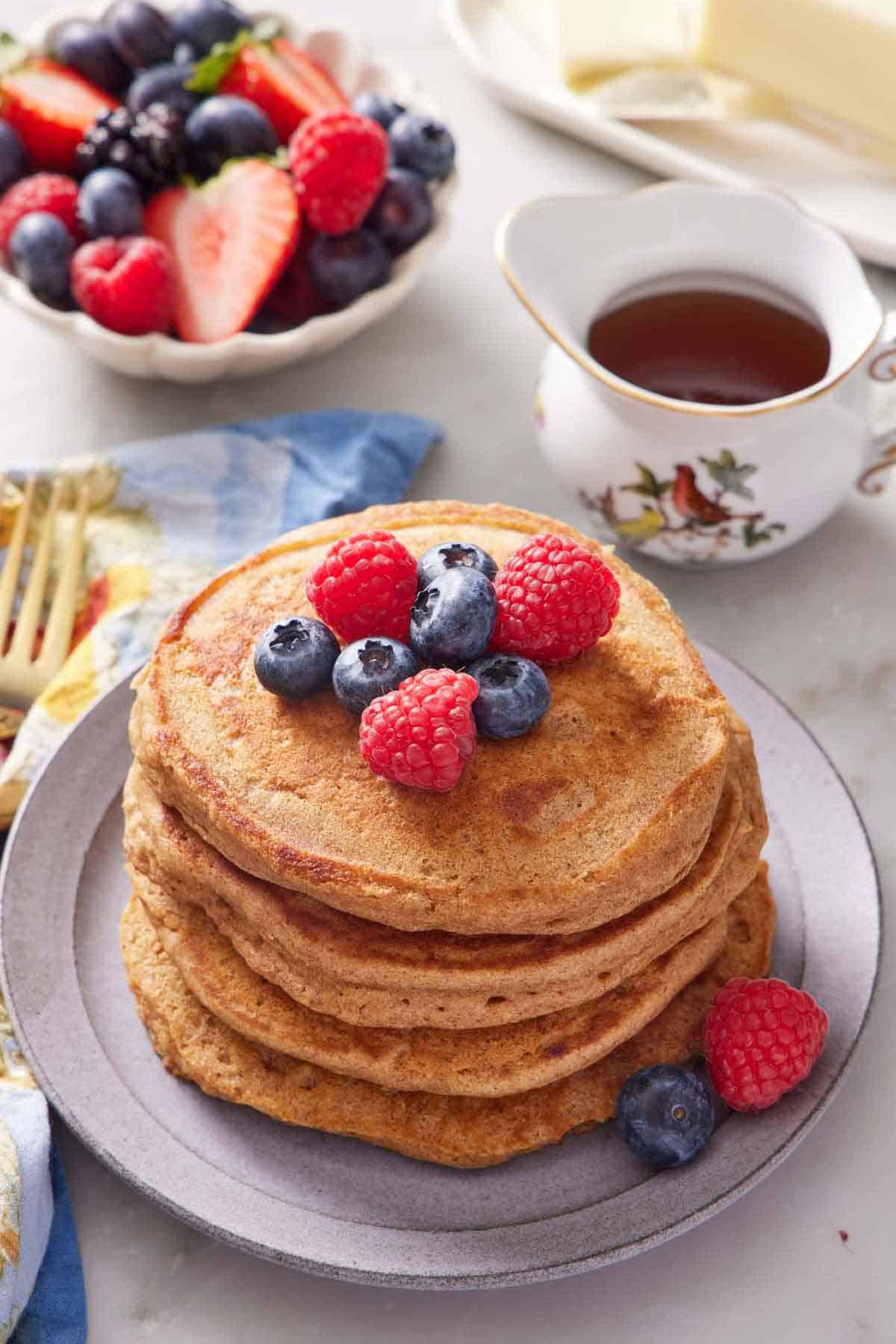 A stack of whole wheat pancakes topped with assorted berries. A bowl of more berries and syrup in the background.