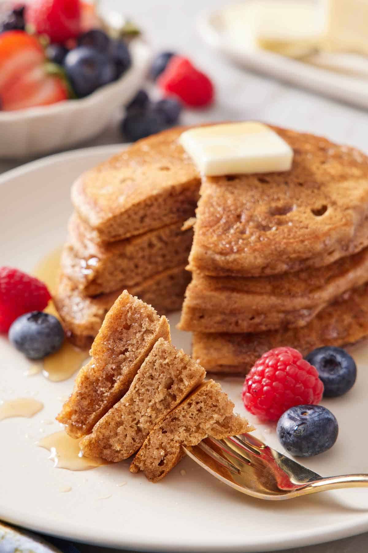 A fork with a bite of whole wheat pancakes on a plate in front of the rest of the pancakes, topped with butter and syrup. Berries scattered around.