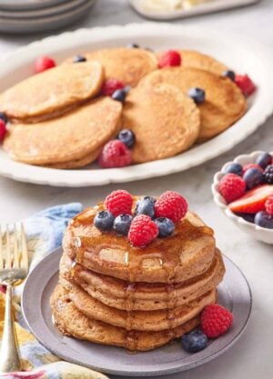 A stack of whole wheat pancakes topped with berries and syrup with a plate of more pancakes in the background along with berries.