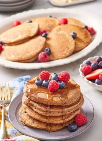 A stack of whole wheat pancakes topped with berries and syrup with a plate of more pancakes in the background along with berries.