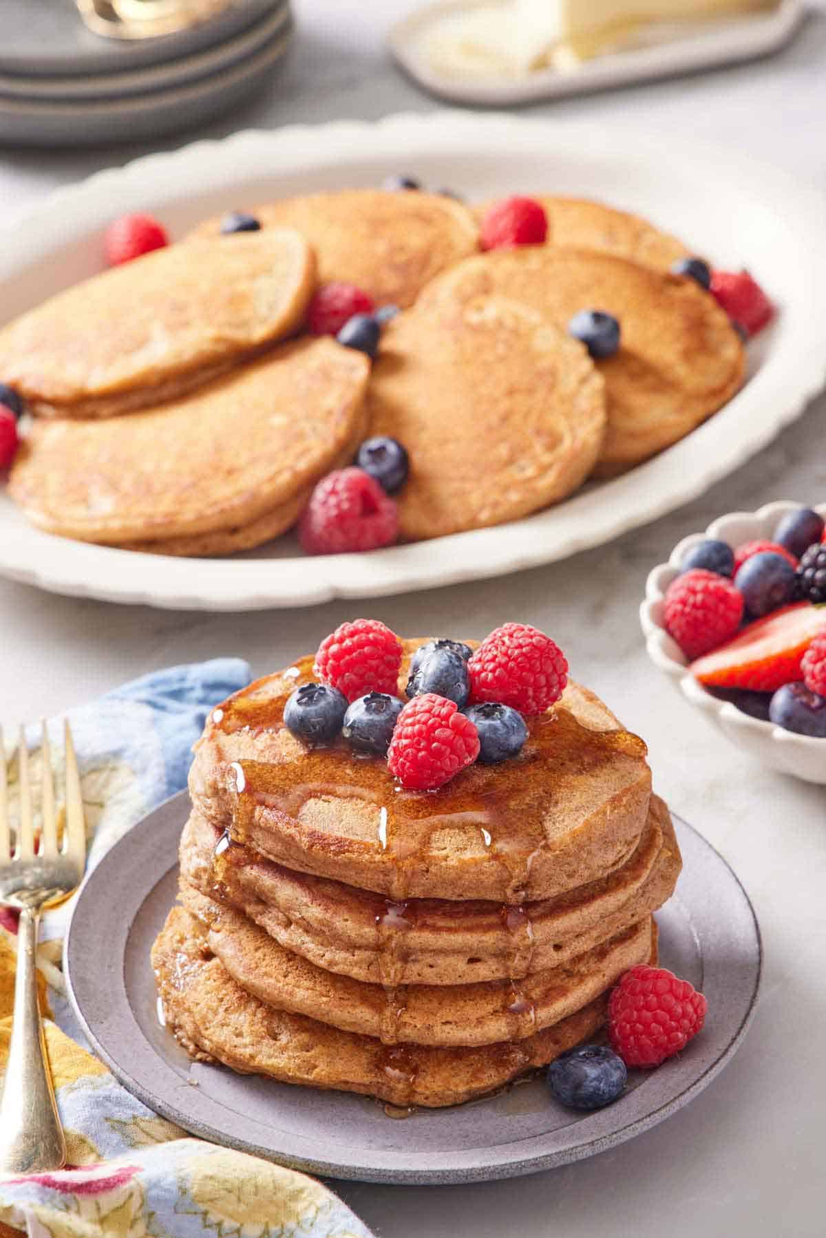 A stack of whole wheat pancakes topped with berries and syrup with a plate of more pancakes in the background along with berries.