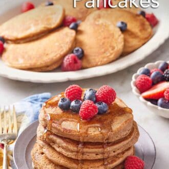 Pinterest graphic of a stack of whole wheat pancakes topped with berries and syrup with a plate of more pancakes in the background along with berries.