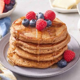 A stack of four whole wheat pancakes topped with berries and syrup. More syrup in the background along with a bowl of berries.