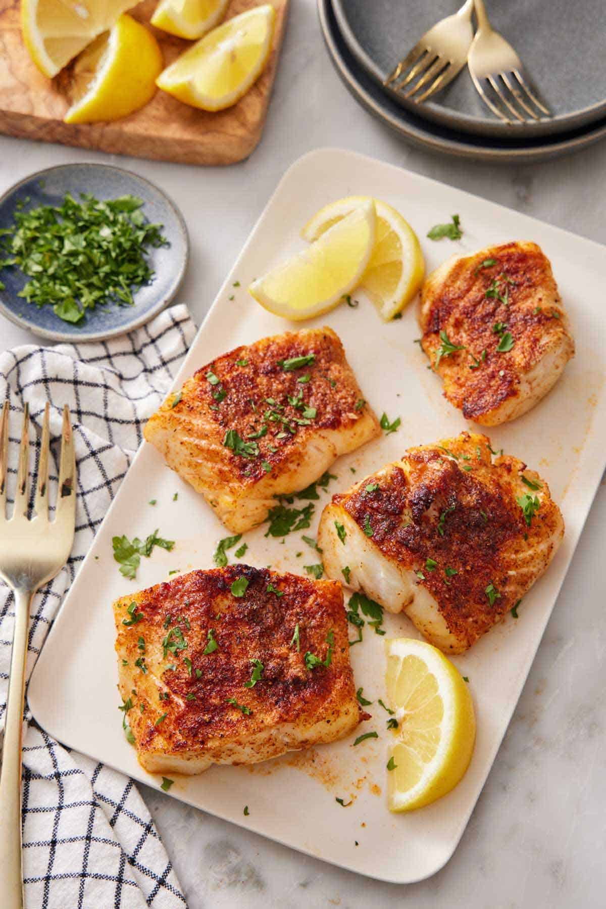 A platter with four pieces of air fryer cod and lemon wedges, garnished with chopped parsley. A bowl of chopped parsley and cut lemon wedges in the background.