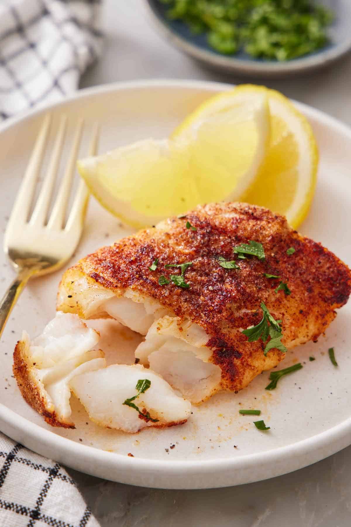 A plate with an air fryer cod with a portion flaked off with two lemon wedges in the background.